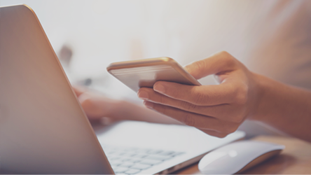 Detail ruky používateľa držiaceho smartfón pri klávesnici, znázorňujúci digitálnu komunikáciu v rámci bankovej aplikácie.